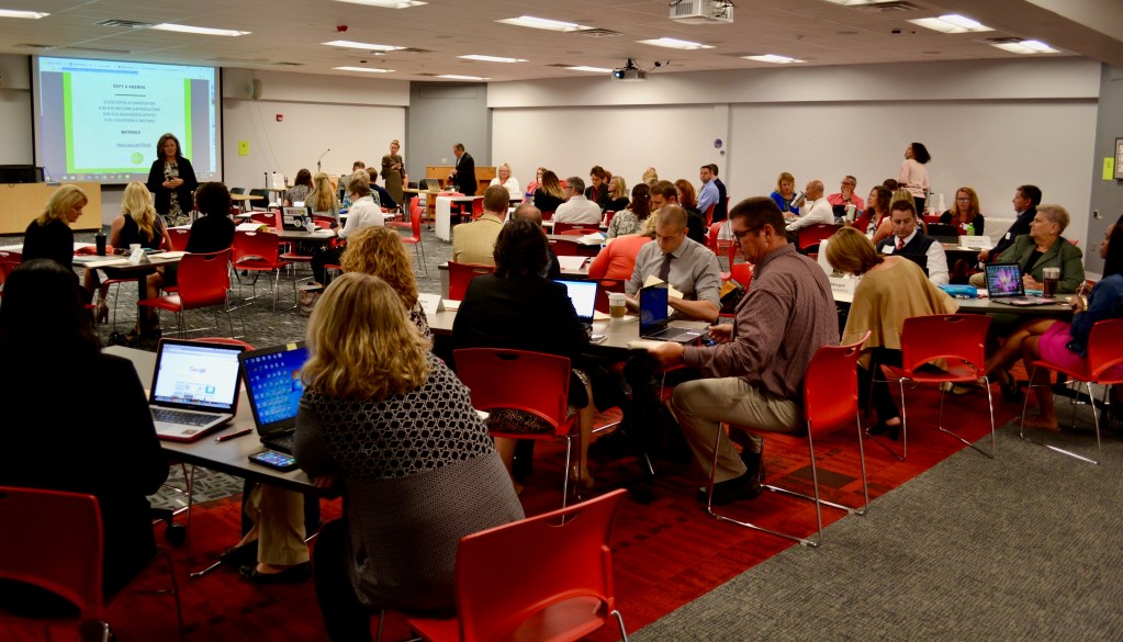 A room full of adults sitting at tables listening to someone speak.